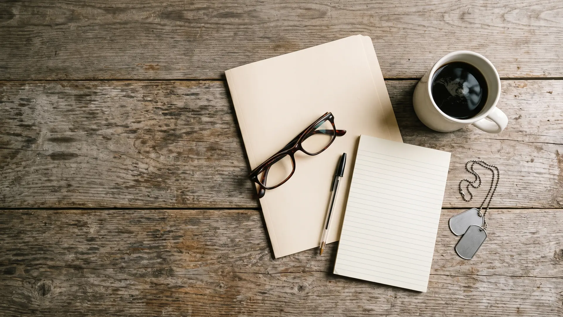 A folder, reading glasses, a pen, a notepad, a mug of coffee and a set of blank dog tags on a wooden table